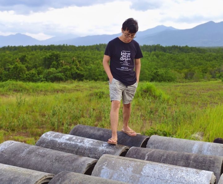 A.B. Nostoria standing on stacked concrete culverts in an open green field outside Puerto Princesa, Palawan, forested hills and mountains behind.